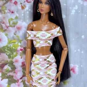 May include: A fashion doll with long black hair and gold earrings, wearing a matching two-piece outfit. The off-the-shoulder crop top and mini skirt feature a pink, white, and gold geometric pattern. The doll has a tan complexion and is posed against a floral backdrop.
