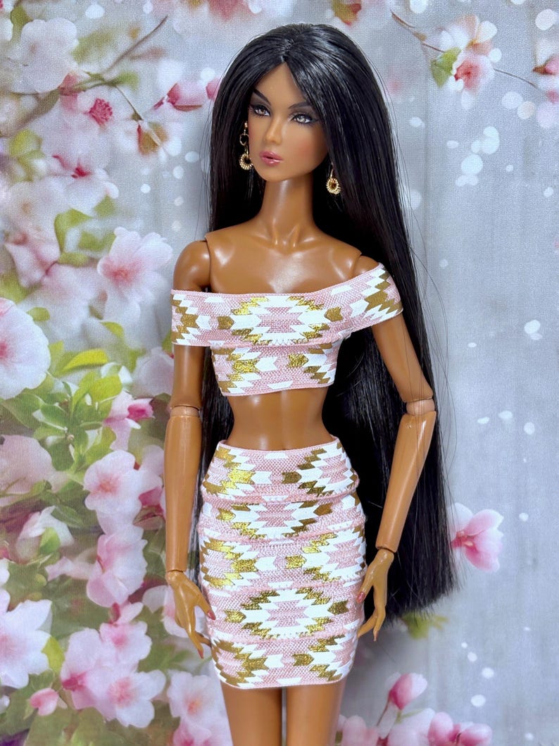 May include: A fashion doll with long black hair and gold earrings, wearing a matching two-piece outfit. The off-the-shoulder crop top and mini skirt feature a pink, white, and gold geometric pattern. The doll has a tan complexion and is posed against a floral backdrop.