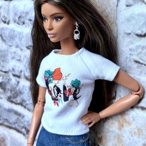 May include: A doll wearing a white t-shirt with a floral and graphic design, paired with blue denim jeans. The doll has long brown hair and is wearing silver earrings. A gold chain belt is visible.