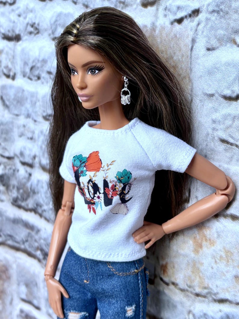 May include: A doll wearing a white t-shirt with a floral and graphic design, paired with blue denim jeans. The doll has long brown hair and is wearing silver earrings. A gold chain belt is visible.