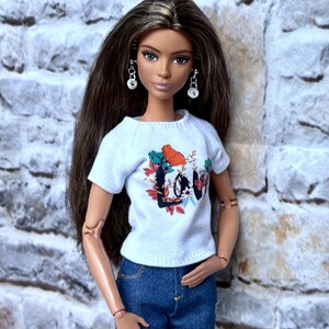 May include: A doll wearing a white t-shirt with a floral and text design that says "Love", blue denim jeans with distressed details, and silver earrings. The doll has long brown hair.