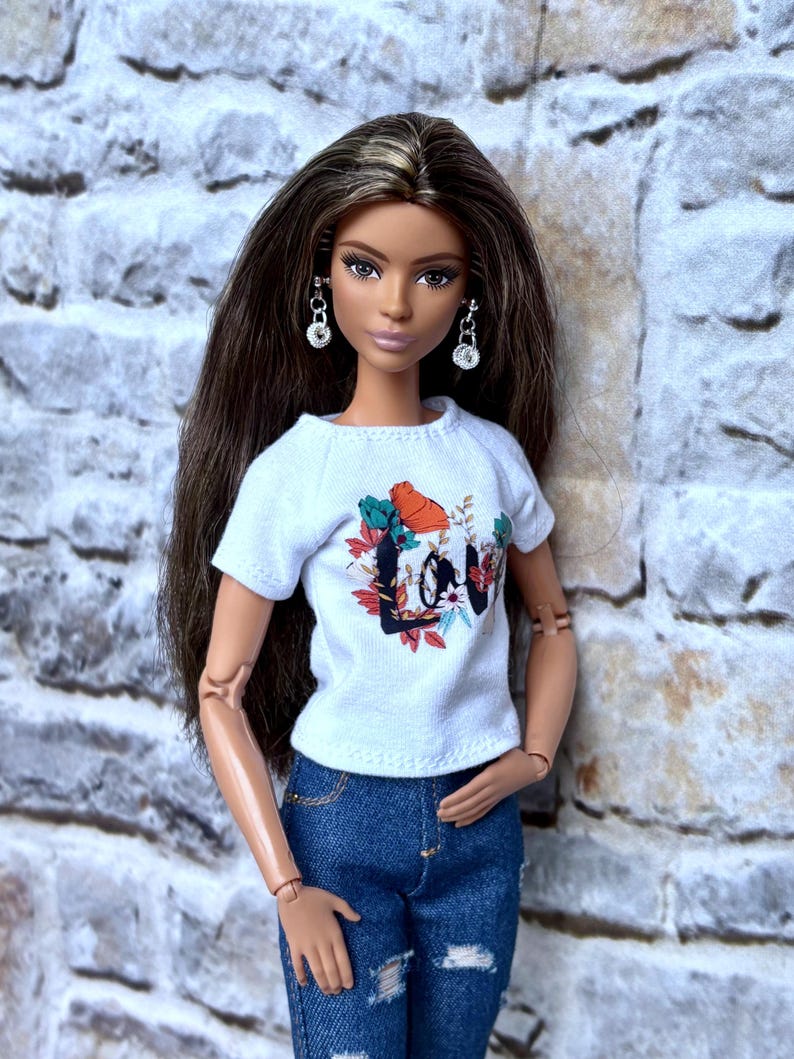 May include: A doll wearing a white t-shirt with a floral and text design that says "Love", blue denim jeans with distressed details, and silver earrings. The doll has long brown hair.