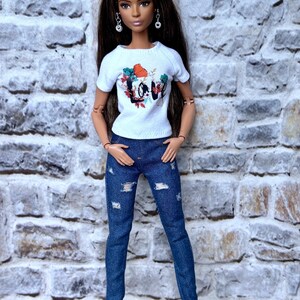 May include: A doll wearing a white t-shirt with a colorful floral and "LOVE" graphic, paired with distressed blue jeans and white sneakers. The doll has long brown hair and is posed in front of a brick wall.