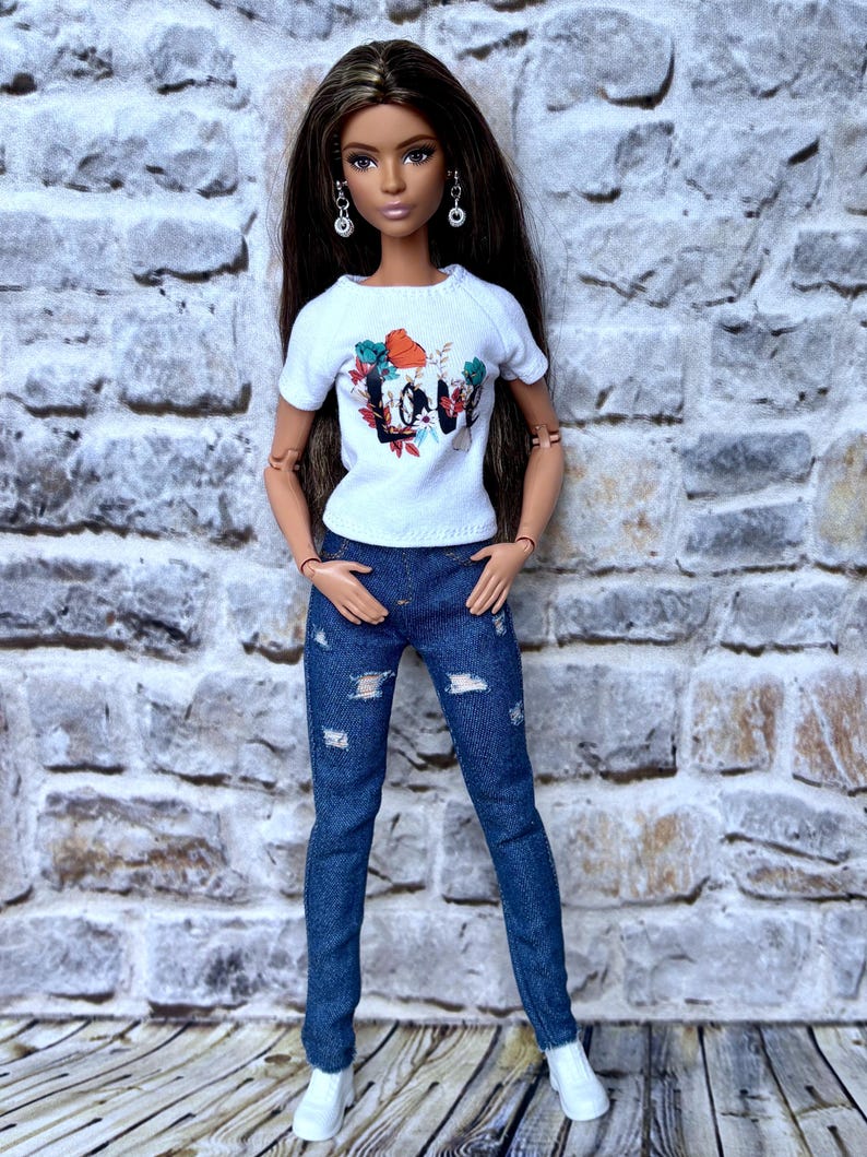 May include: A doll wearing a white t-shirt with a colorful floral and "LOVE" graphic, paired with distressed blue jeans and white sneakers. The doll has long brown hair and is posed in front of a brick wall.
