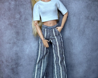 May include: A fashion doll with long blonde hair, wearing a white crop top and wide-leg pants with vertical white stripes on a dark blue background. The doll is also wearing silver hoop earrings and silver shoes.