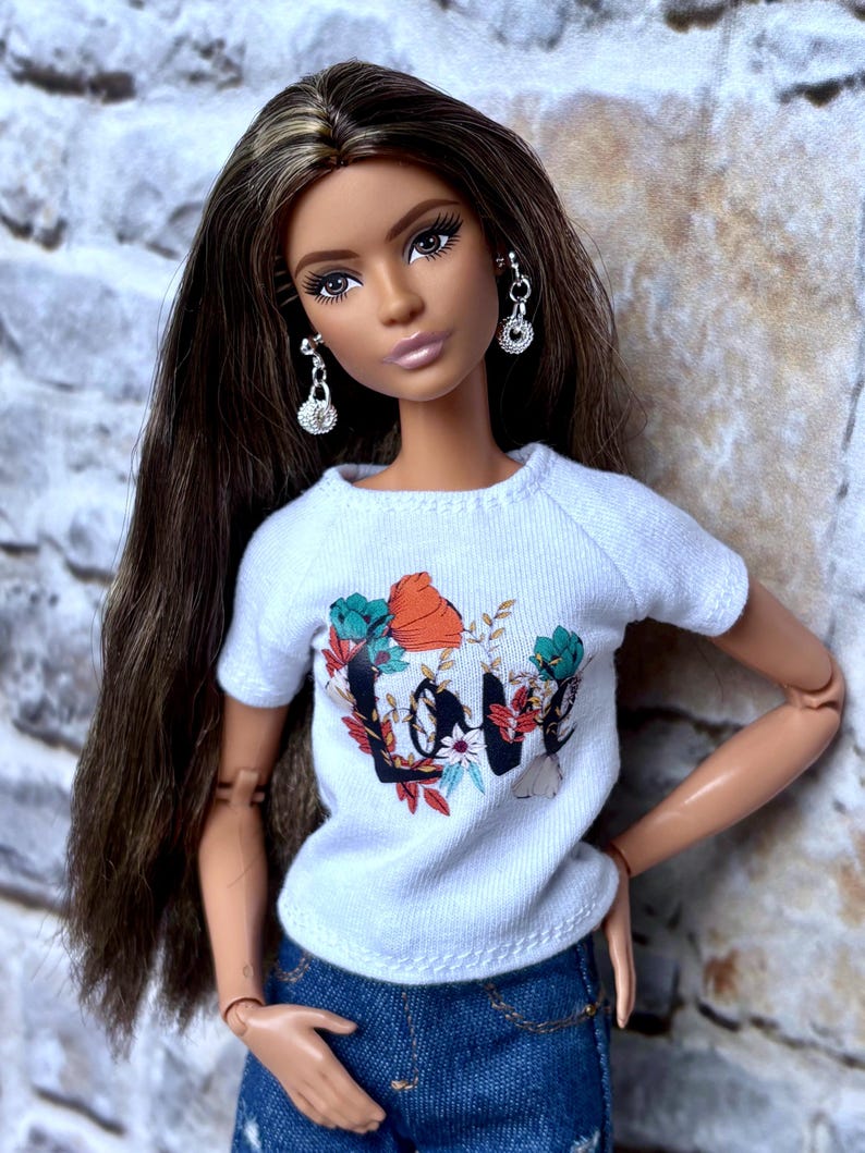 May include: A doll with long brown hair and silver earrings. The doll wears a white t-shirt with a floral "Love" graphic and blue denim jeans. The doll is posed with one arm bent at the elbow and the other arm at the side.