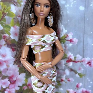 May include: A fashion doll with long brown hair and dangling earrings. The doll wears a two-piece outfit with a pink, white, and gold geometric pattern. The off-the-shoulder crop top and matching skirt create a stylish ensemble. The doll is posed in front of a floral background.
