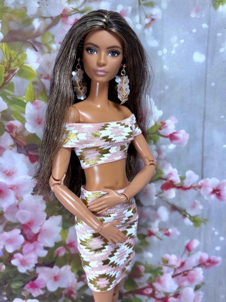 May include: A fashion doll with long brown hair and dangling earrings. The doll wears a two-piece outfit with a pink, white, and gold geometric pattern. The off-the-shoulder crop top and matching skirt create a stylish ensemble. The doll is posed in front of a floral background.