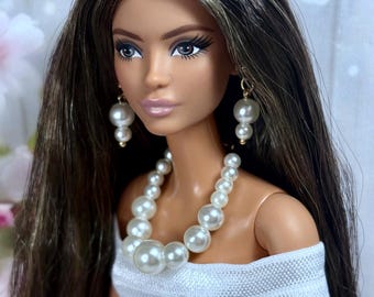 May include: A doll with long brown hair and pearl jewelry, including a necklace and earrings. The doll is wearing a white off-the-shoulder top. The doll has makeup on, including eye shadow and lipstick. The background is blurred with pink and white floral elements.