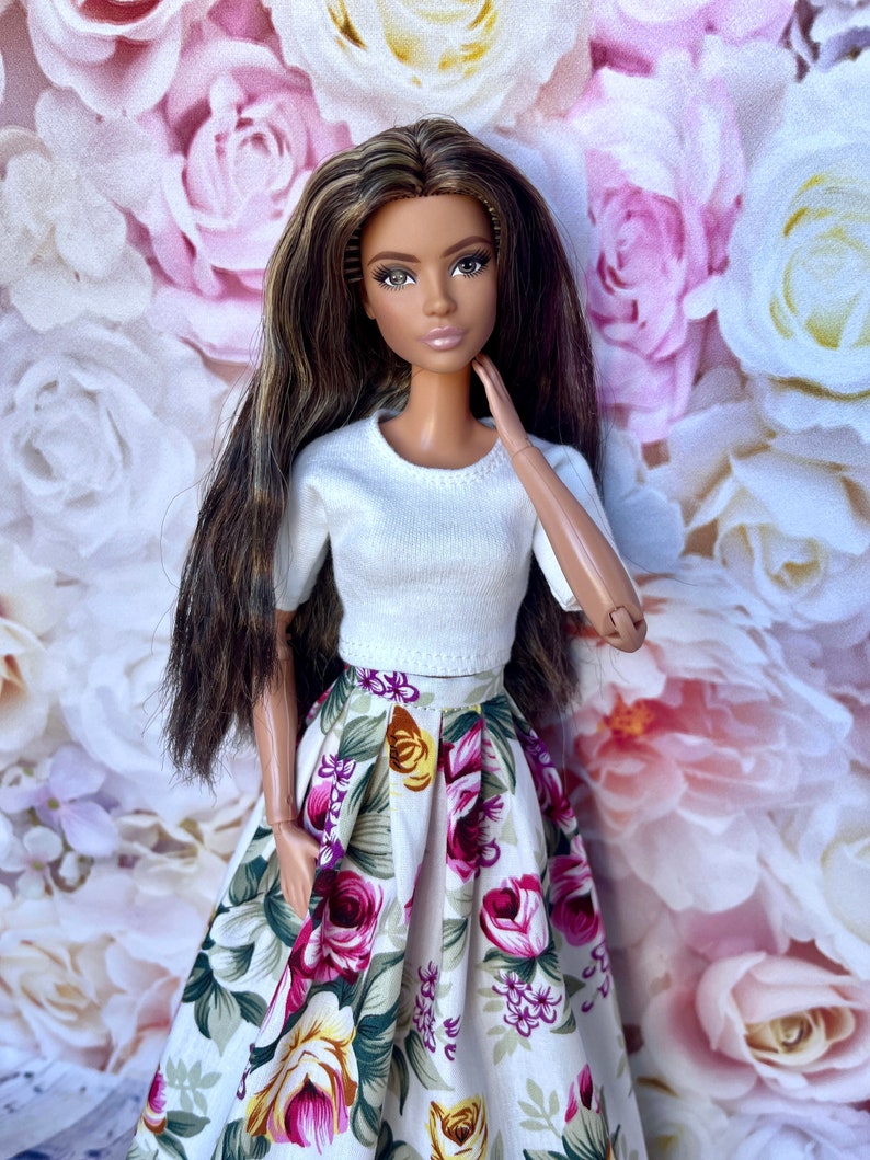 Peut inclure: Poup&eacute;e aux longs cheveux bruns, v&ecirc;tue d'un haut blanc &agrave; manches courtes et d'une jupe &agrave; imprim&eacute; floral. La jupe pr&eacute;sente un motif de fleurs roses, jaunes et vertes sur fond blanc. La poup&eacute;e est pos&eacute;e devant une toile de fond florale.