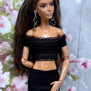 May include: A doll in a black off-the-shoulder crop top and matching mini skirt. The doll has long brown hair and silver earrings. The outfit has a scalloped trim. The background features pink and white flowers.