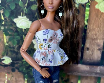 May include: A doll with long brown hair wearing a white floral top and blue jeans. She has brown eyes and is wearing square earrings.