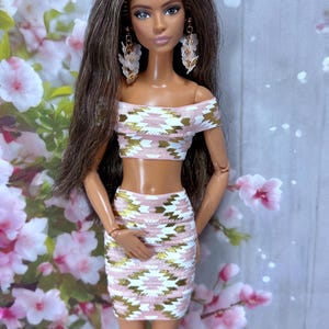 May include: A doll wearing a pink, white, and gold patterned crop top and matching skirt. The doll has long brown hair and is wearing floral earrings. The background features pink flowers.