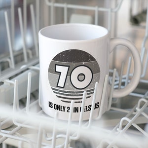 A white 11-ounce ceramic mug with a comfortable C-handle and rounded edges. The black and white design displays the words 70 Is Only 21 in Celsius.