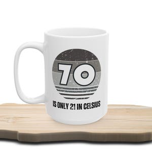 White ceramic mug on a wooden surface, featuring a vintage-style circular graphic with the number “70” and the text “Is only 21 in Celsius” in black lettering.