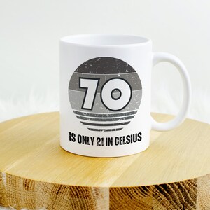 White ceramic mug on a round wooden surface, featuring a vintage-style circular graphic with the number “70” and the text “Is only 21 in Celsius” in black lettering.
