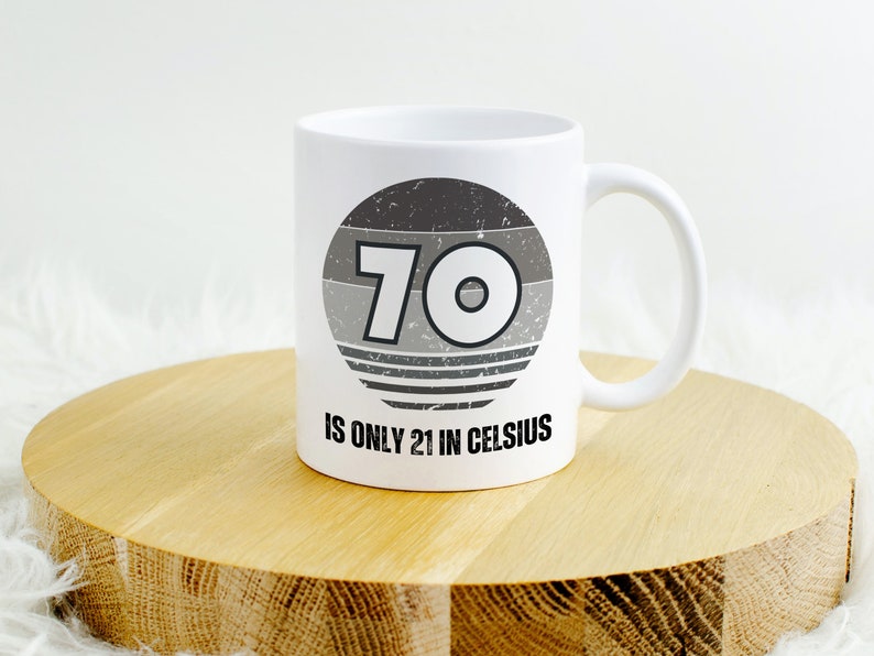 White ceramic mug on a round wooden surface, featuring a vintage-style circular graphic with the number “70” and the text “Is only 21 in Celsius” in black lettering.