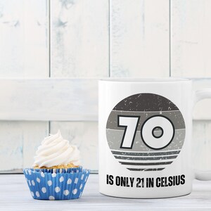 A white 11-ounce ceramic mug with a comfortable C-handle and rounded edges. The black and white design displays the words 70 Is Only 21 in Celsius.