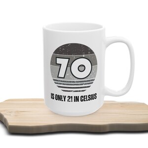 White ceramic mug on a wooden surface, featuring a vintage-style circular graphic with the number “70” and the text “Is only 21 in Celsius” in black lettering.