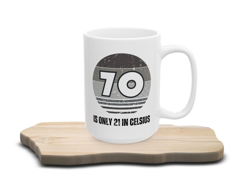 White ceramic mug on a wooden surface, featuring a vintage-style circular graphic with the number “70” and the text “Is only 21 in Celsius” in black lettering.