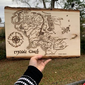 May include: A wooden plaque with a map of Middle Earth, featuring intricate engraved details. The map includes geographical features and text, with the words "Middle Earth" at the bottom. The plaque has a natural bark edge.