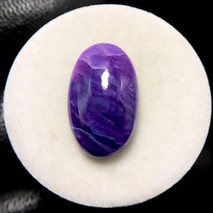 May include: An oval-shaped, polished gemstone with a deep purple hue, exhibiting lighter streaks and patterns throughout. The stone is set against a white, circular backdrop, highlighting its color and texture. The gemstone's surface reflects light, showcasing its glossy finish.