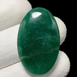 May include: Oval-shaped, polished green gemstone with a smooth, glossy surface. The stone has a speckled appearance and a subtle, light-colored vein running through it. The gemstone is held against a white fabric background.