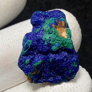 Può includere: Primo piano di un campione minerale grezzo di azzurrite. La pietra presenta un colore blu intenso con macchie verdi e un tocco di marrone. La superficie ha un aspetto ruvido e strutturato, che mostra la formazione naturale del minerale.