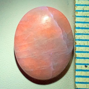 May include: Oval-shaped, polished gemstone with a peach and pink color gradient. The stone has a smooth, reflective surface with subtle internal striations. A ruler with blue and yellow markings is visible on the right side.