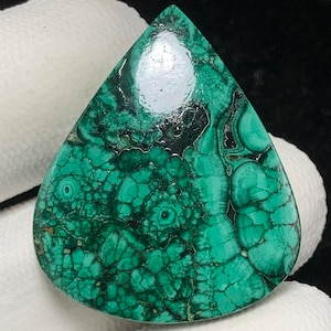 Puede incluir: Una piedra preciosa de malaquita en forma de lágrima con una superficie pulida. La piedra presenta un color verde vibrante con intrincados patrones de verde más oscuro y negro, creando un diseño orgánico único. La piedra preciosa se utiliza probablemente para joyería o fines decorativos.