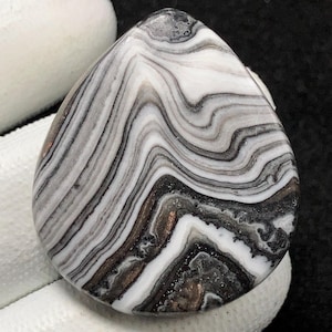 Puede incluir: Un cabujón de ágata blanco y negro con un patrón de remolino. La piedra tiene un acabado natural y pulido y es un hermoso ejemplo de este tipo de piedra preciosa.