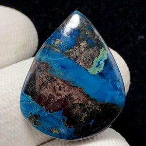 May include: A polished, teardrop-shaped gemstone with a vibrant mix of colors. The stone features a deep blue base, accented with streaks of brown, black, and hints of green. The surface is smooth and reflective, showcasing the natural patterns.
