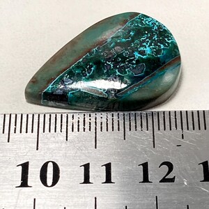 May include: A polished, teardrop-shaped gemstone with a mix of teal, dark green, and brown hues. The stone has a unique, textured pattern and is placed next to a ruler for size reference.