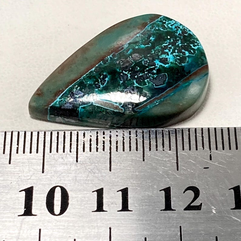 May include: A polished, teardrop-shaped gemstone with a mix of teal, dark green, and brown hues. The stone has a unique, textured pattern and is placed next to a ruler for size reference.