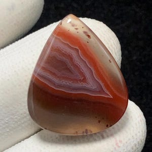 May include: A teardrop-shaped agate cabochon with a red, white, and brown banded pattern. The stone has a smooth, polished surface.