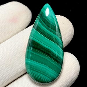 May include: A polished, teardrop-shaped malachite gemstone. The stone features bands of varying shades of green, creating a striped pattern. The gemstone is held by a white gloved hand, with a black background.