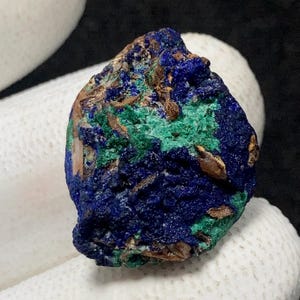 Puede incluir: Primer plano de un espécimen mineral de azurita en bruto. La piedra presenta un color azul profundo con parches de inclusiones verdes y marrones vibrantes. El mineral se sostiene sobre un fondo blanco.