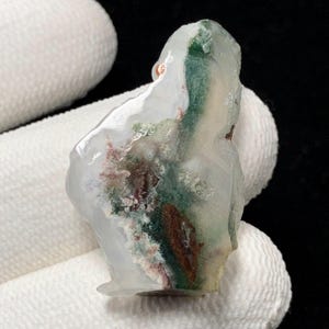 May include: A translucent, irregularly shaped gemstone with internal patterns of green, brown, and red. The stone has a clear, glassy appearance with a rough, natural texture. The colors are swirled and layered within the stone, creating a unique, organic design.