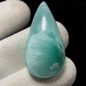 Puede incluir: Un cabujón de magnesita verde y blanca en forma de lágrima con un patrón de remolino. La piedra está pulida y tiene un acabado liso y brillante.
