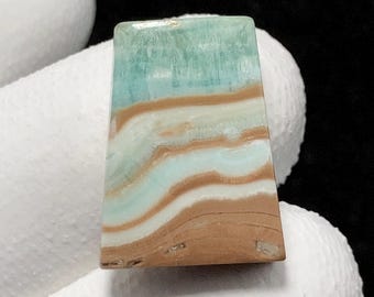 May include: A rectangular gemstone with layers of light blue, white, and brown. The top is light blue, transitioning to white and brown bands. The stone is held by a gloved hand. The gemstone's layers are reminiscent of a landscape.