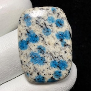 May include: A polished K2 stone with a rectangular shape and rounded corners. The stone features a creamy white base with vibrant blue orb-like patterns and black veining throughout. This unique gemstone is ideal for jewelry making or collecting.