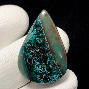 May include: A polished, teardrop-shaped gemstone. It displays a blend of teal, turquoise, and brown colours, with dark maroon inclusions. The gemstone is held against a white fabric background.