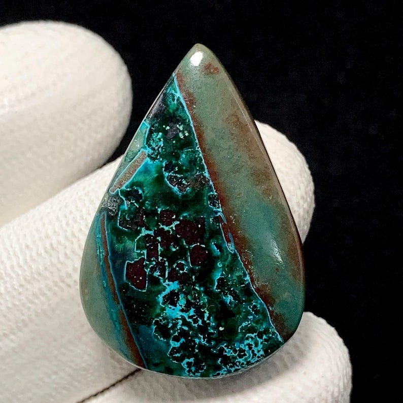 May include: A teardrop-shaped gemstone with a polished surface. The stone features a mix of teal, turquoise, and brown hues, with dark maroon inclusions. The gemstone is held against a white fabric background.