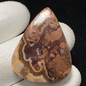 Könnte beinhalten: Ein tropfenförmiger Edelstein mit polierter Oberfläche. Der Stein weist eine Mischung aus Farben auf, darunter Brauntöne, Kupfer, Creme und Hauch von Orange. Das Muster hat ein geschichtetes, wirbelndes Aussehen mit einer polierten, reflektierenden Oberfläche.