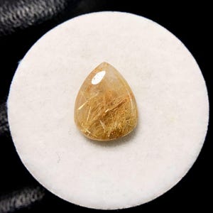 May include: A teardrop-shaped gemstone with a golden-brown hue, featuring internal needle-like inclusions. The gem is set against a white, circular backdrop, with a dark background.