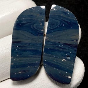 May include: Two polished, semi-circular stones with a deep blue, swirling pattern resembling a night sky. The stones have white speckles and a glossy finish, ideal for jewelry making or crafting.
