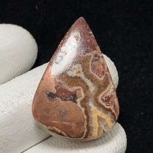 May include: A teardrop-shaped gemstone with a polished surface. The stone features a mix of colors, including copper, brown, white, and hints of yellow. The pattern resembles natural formations. The gemstone is held by a gloved hand against a dark background.