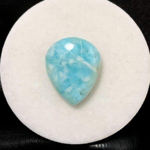Puede incluir: Una piedra preciosa cabujón de color azul claro con vetas blancas. La piedra preciosa tiene forma de lágrima y una superficie lisa y pulida.