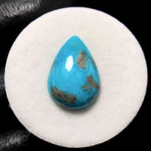 May include: A teardrop-shaped turquoise gemstone with dark brown matrix patterns. The polished stone is set on a white circular surface, with a black background. The gemstone is a vibrant blue color.