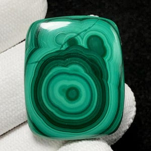 May include: A polished, rectangular malachite stone with a vibrant green color and concentric patterns. The stone's surface displays a swirling, eye-like design, showcasing varying shades of green.